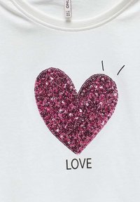 T-shirt en coton blanc avec un grand cœur en paillettes roses et le mot "LOVE" en dessous en lettres noires en gras.