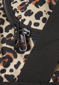 Pungulvogn i leopardmønster med beige bakgrunn, med svarte og brune flekker; inkluderer sorte glidelåsgrep og stropp. Teksturert stoffoverflate.