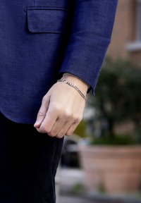 Acercamiento de una mano con una pulsera de cadena de plata, vistiendo una chaqueta azul y pantalones negros, al aire libre con fondo desenfocado.