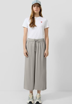 Młoda kobieta w jasnoniebieskiej czapce, białej koszulce z małym czerwonym sercem, szerokich szarych spodniach i beżowych trampkach stoi na jednolitym tle.