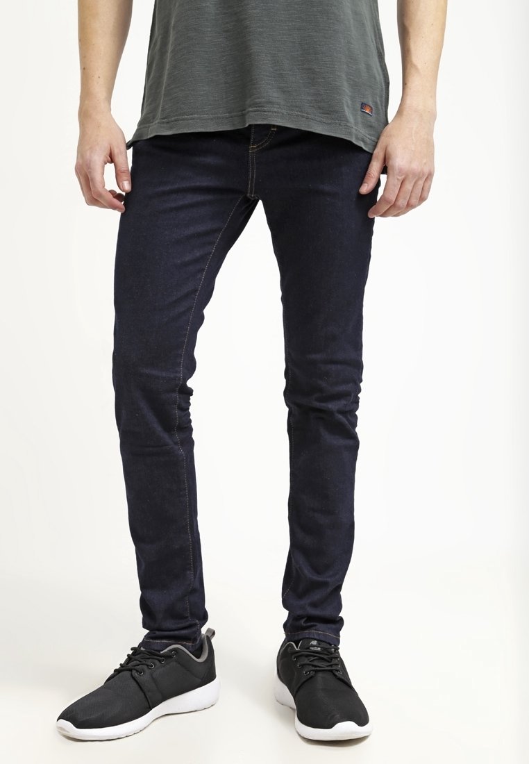 Vaqueros pitillo de mezclilla azul marino con textura suave y costuras en contraste, combinados con zapatillas deportivas negras con suela blanca.