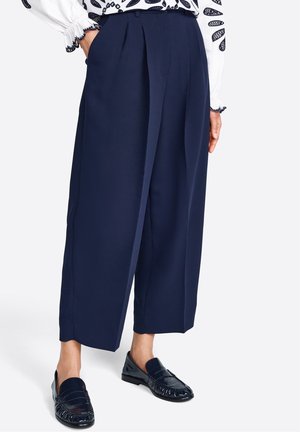 Granatowe szerokie spodnie typu cropped zestawione z czarnymi lakierowanymi loafersami i białą bluzką z czarnymi wzorami liści.