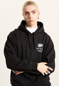 Black hooded sweatshirt made of cotton-blend fabric. Features a front logo print in white. Model is wearing rings and earrings.