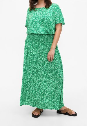Femme portant un haut vert à manches courtes et une jupe longue assortie à taches blanches, associée à des sandales marron à brides croisées.
