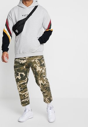 Felpa grigia con cappuccio e maniche a strisce nere e colorate. Pantaloni cargo mimetici e sneakers bianche. Borsa a tracolla nera.