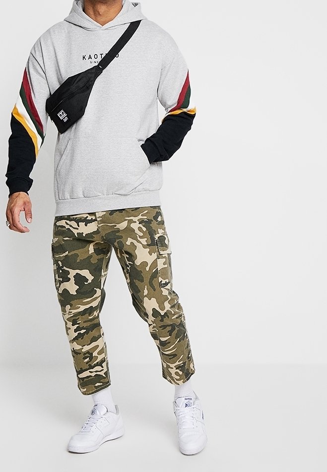 Felpa grigia con cappuccio e maniche a strisce nere e colorate. Pantaloni cargo mimetici e sneakers bianche. Borsa a tracolla nera.