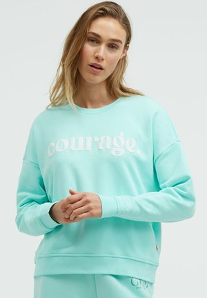 Junge Frau in hellblauem Sweatshirt und passender Hose, auf dem Sweatshirt steht "Mut." Die Hände sind sanft voreinander gelegt.