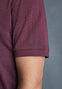Bordo majica s teksturiranim materijalom, koja ima rebrasti okrugli izrez i kratke rukave. Bočni pregled koji prikazuje detalje rukava i ruke.