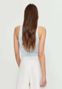 Vrouw met lang bruin haar, gekleed in een lichtblauwe mouwloze top en hoge witte broeken, staande met de rug naar de camera tegen een effen achtergrond.