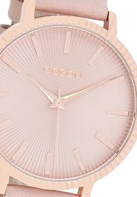 Montre en or rose avec un cadran rose texturé, des marqueurs d'heure nets et des aiguilles minimalistes. Le boîtier présente un bord nervuré. Bracelet en cuir doux.