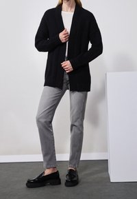 Svart stickad cardigan med en texturerad tyg, öppen framtill och ribbade kanter, kombinerad med grå jeans och svarta slip-on skor.