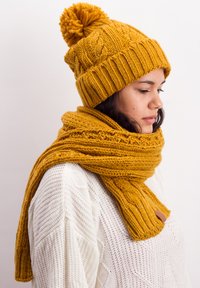Mustard yellow knit beanie with pom-pom and matching chunky scarf featuring textured patterns, worn over a white, textured sweater.