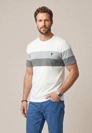 Homme avec barbe portant un t-shirt rayé blanc et gris et un jean bleu, debout avec une main dans la poche sur un fond uni.