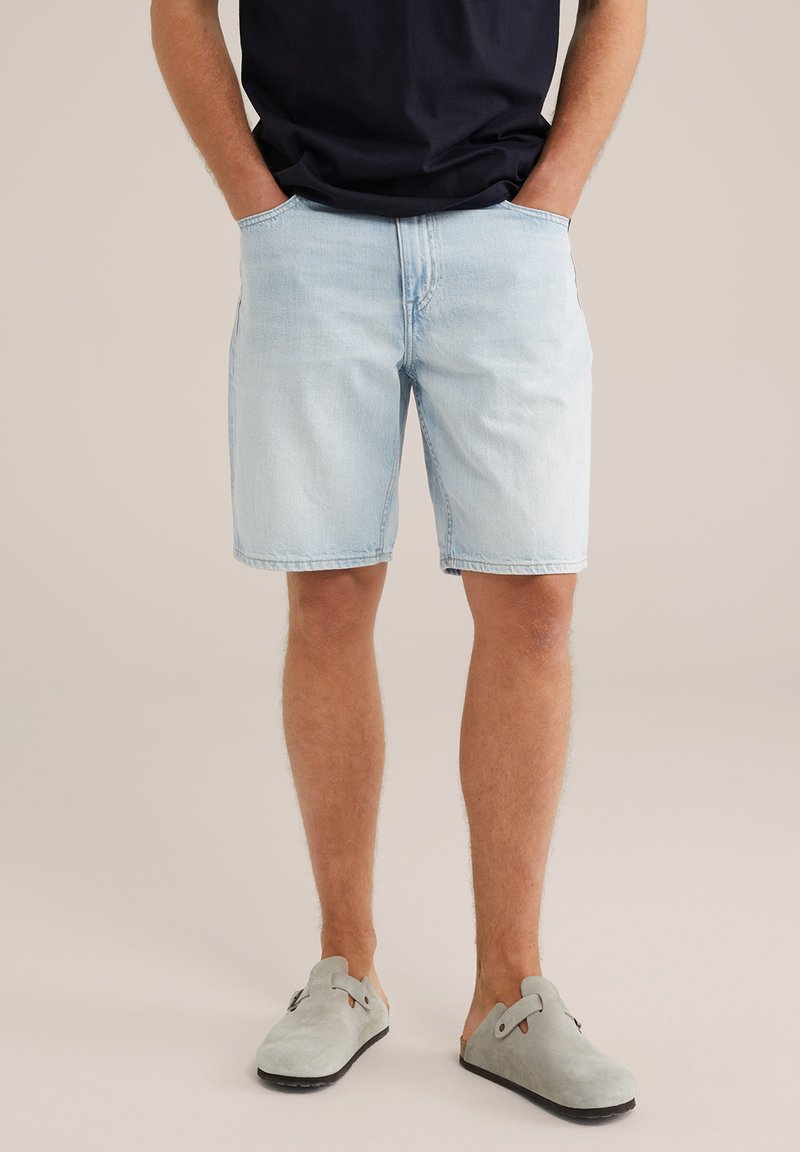 Gai zils denima šorti ar taisnu griezumu, ar sānu kabatām un klasisku jostu. Valkāti ar pelēkiem slip-on apaviem.