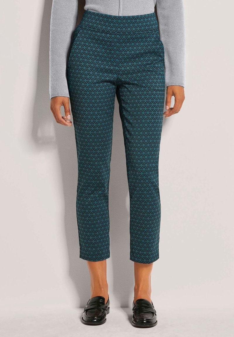 Femme portant un pantalon slim turquoise avec un motif géométrique, un haut gris à manches longues et des mocassins en cuir noirs, debout contre un mur clair.
