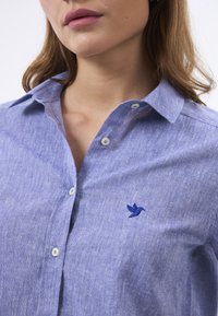 Camicia button-up azzurro chiaro realizzata in tessuto testurizzato, con un logo di uccello ricamato sul petto e colletto morbido e arrotondato.