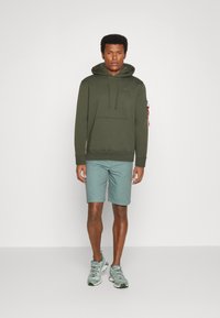 Felpa con cappuccio verde oliva con tasca frontale, abbinata a pantaloni corti turchesi chiari e sneakers grigie. Presenta un design minimalista e dettagli eleganti.