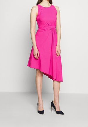 Kobieta ubrana w różową sukienkę bez rękawów z asymetrycznym dołem i dopasowanym paskiem w talii, w połączeniu z czarnymi butami na wysokim obcasie.