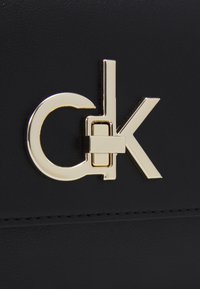 Černý syntetický materiál s kovovým logem ve zlaté barvě, které kombinuje písmena 'C', 'K' a kulatý prvek v lesklém provedení.