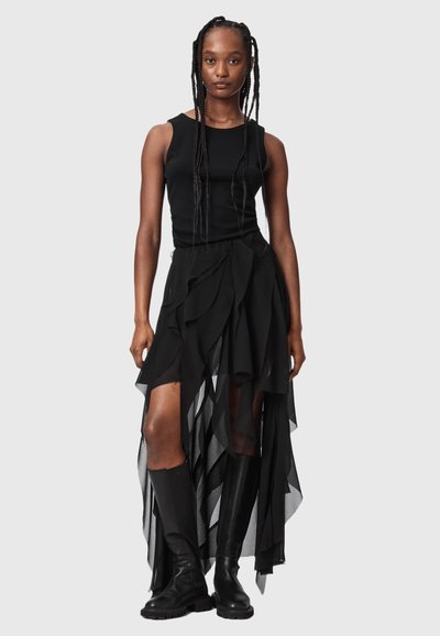Femme avec de longues tresses portant un haut noir sans manches, une jupe noire asymétrique à plusieurs couches, et des bottes noires montantes jusqu'aux genoux, debout devant un fond uni.