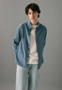 Blå skjorta med krage och ficka. Buren över en ljust grå t-shirt och kombinerad med ljusa jeans. Slät textur.