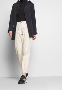 Giacca leggera nera con cappuccio e zip, abbinata a pantaloni color crema con polsini elastici e scarpe sportive nere. Design semplice e funzionale.