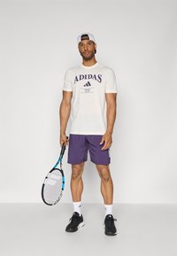 Hvit T-skjorte med korte ermer og lilla Adidas-logo, kombinert med lilla shorts og svarte tennissko. Holder en tenniskølle.