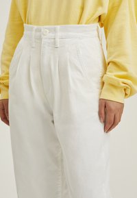 Pantalones de algodón blancos con pliegues y talle alto, que cuentan con bolsillos laterales y cierre de botón, combinados con una blusa de manga larga amarilla.