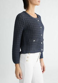 Marine gebreide cardigan met een textuurpatroon, ronde halslijn en vijf metalen knopen aan de voorkant, gecombineerd met witte high-waisted broeken.