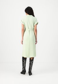 Rochie călcată verde deschis cu mâneci suflecate, talie strânsă și un croi până la genunchi. Asortată cu cizme negre până la genunchi cu broderie argintie.
