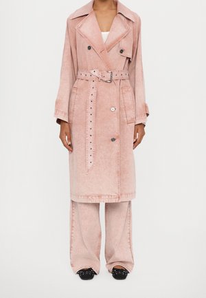 Femme portant un long trench-coat rose avec ceinture, un pantalon large et des chaussures noires cloutées, debout devant un fond blanc uni.