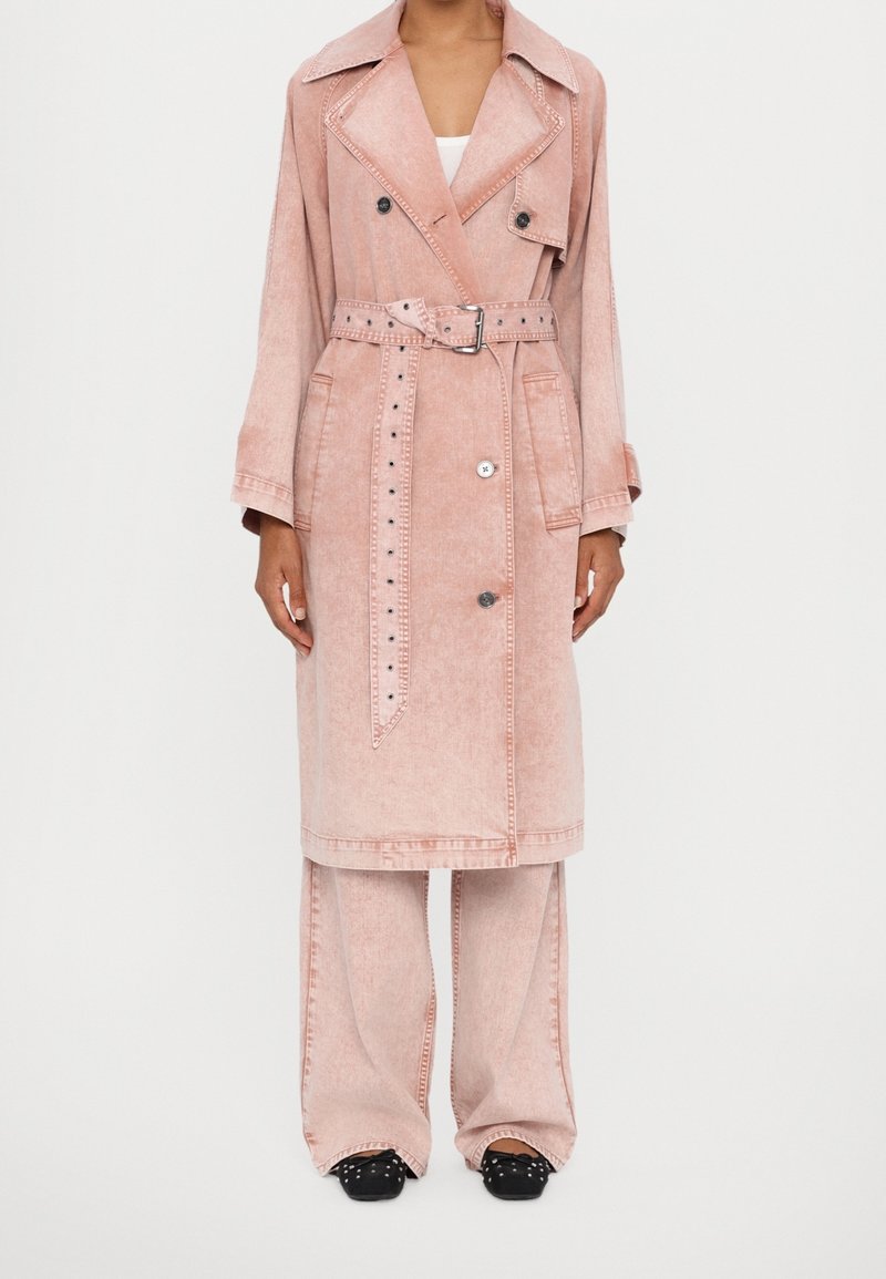 Femme portant un long trench-coat rose avec ceinture, un pantalon large et des chaussures noires cloutées, debout devant un fond blanc uni.
