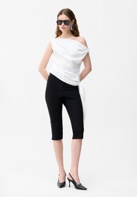 Frau trägt weißes asymmetrisches ärmelloses Oberteil, schwarze Hosen bis zur Mitte der Wade, schwarze High Heels, übergroße Ohrringe und dunkle Sonnenbrille, steht.