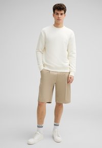 Felpa beige chiaro con polsini e fondo a coste, abbinata a pantaloni corti marrone chiaro. Il modello indossa sneakers bianche e calzini a righe.