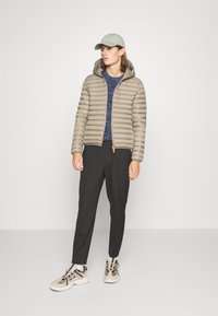 Puffer jacket in light beige with a hood and vertical quilted pattern, paired with a navy top, black trousers, and beige athletic shoes.