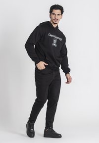 Felpa nera con stampa grafica, materiale in cotone, tasca a marsupio, abbinata a jeans neri e sneakers nere. Modello in posa verticale.