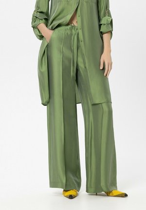 Woman wearing loose green satin pants and matching long shirt, paired with yellow and black pointed high heels.