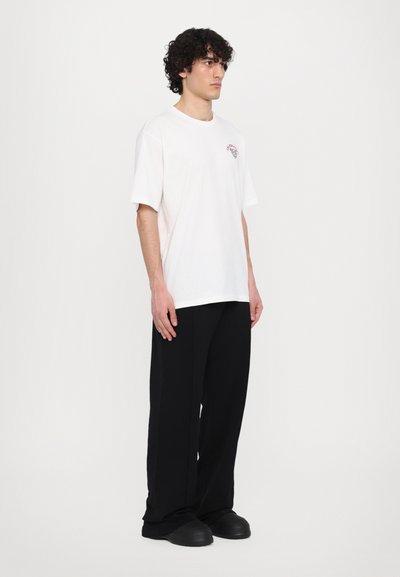 T-shirt en coton blanc à manches courtes et col rond, avec un petit design graphique rouge. Assorti à un pantalon noir ample et des chaussures noires.