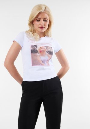 Blondynka stoi z rękami za plecami, ubrana w białą koszulkę z uśmiechniętą kobietą z różowym kwiatem we włosach, w zestawie z czarnymi spodniami.