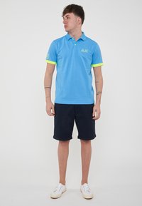 Polo shirt azzurro chiaro con maniche corte, dettagli verde neon sui polsini e logo sulla manica sinistra. Indossato con pantaloni corti blu navy e sneakers bianche.