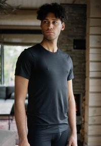 Sort kortærmet t-shirt med en glat tekstur og rund halsudskæring, båret af en model, der står indendørs med træ- og stenharmonier i baggrunden.