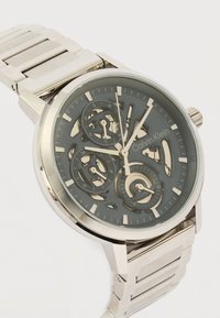 Ασημένιο ρολόι χειρός Calvin Klein με διαφανές καντράν, τρία υποκαντράν και μεταλλικό μπρασελέ, που δείχνει τους εσωτερικούς μηχανισμούς και την ώρα 12:10:57.