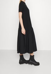 Schwarzes, kurzärmeliges Kleid mit einem gerafften Saum. Hergestellt aus weichem Stoff, bietet es eine lockere Passform. Kombiniert mit klobigen schwarzen Stiefeletten.