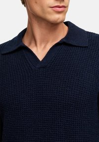 Maglione lavorato a maglia navy con un motivo testurizzato, scollo a V e colletto. Il materiale sembra morbido e leggermente a coste con una vestibilità aderente.