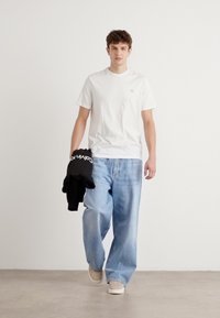 Weißes Baumwoll-T-Shirt, lockere blaue Jeans, hält einen schwarzen Sweatshirt, beigefarbene Sneakers, drinnen mit neutralem Hintergrund.