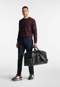 Svart läder weekendväska med strukturerad yta, dragkedjefickor och dubbla handtag. Modellen bär en geometriskt mönstrad tröja och marinblå byxor.