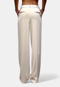 Pantaloni larghi crema con una texture liscia, dotati di due tasche posteriori e una vestibilità su misura. La cintura è progettata per un look a vita bassa.