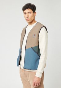 FAGUO fleece vest met een beige en blauwe kleurblok ontwerp, rits aan de voorkant en een ronde logo op de borst. Zacht, gestructureerd materiaal.
