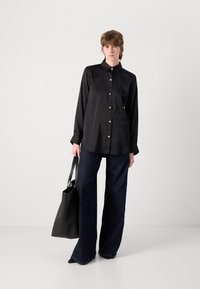 Camisa preta de manga longa com colarinho e botões dourados, emparelhada com calças largas de denim escuro. Uma mala tote preta repousa na mão da modelo.