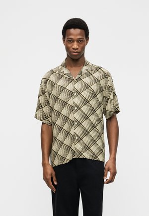 Muž v béžovej a čierno-kockovanej krátkorukáve košeli a čiernych nohaviciach stojí pred jednoduchým svetlým pozadím.